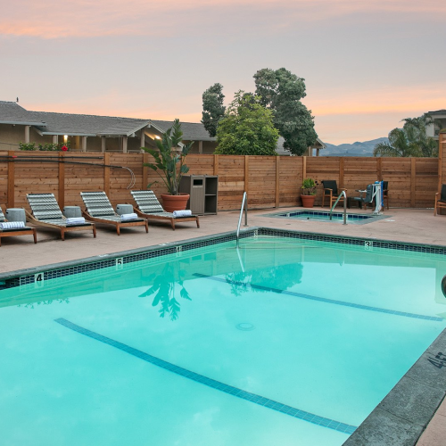 A calm backyard pool with turquoise water, lounge chairs along the deck, a wooden fence, and a small dining setup under a gazebo at sunset.