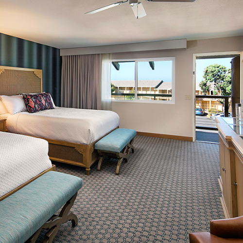 Two double beds with white linens, teal benches, a desk area, flat-screen TV, and a balcony view of a sunny hotel suite with blue accents, ending now.
