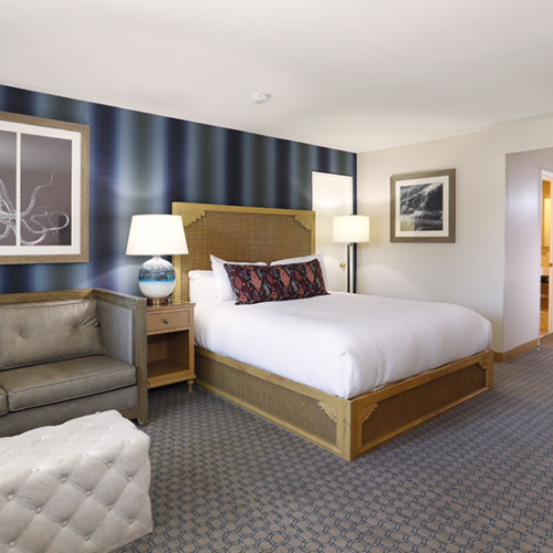 A hotel room with a wooden bed, two lamps, a navy accent wall with framed art, a sofa, and a small entry hallway, cozy and modern.