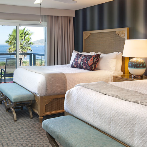 Two queen beds in a seaside hotel room with a balcony, nightstands, lamps, and a view of the ocean and palm trees outside.