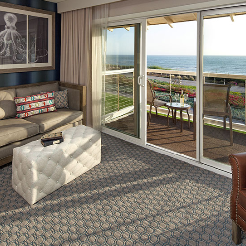 A cozy hotel living area with a beige sofa, tufted ottoman, ocean-view balcony through sliding glass doors, and a patterned carpet.