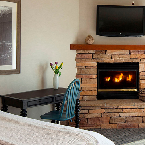 A cozy hotel room with a stone fireplace, lit fire, mounted TV, wooden mantel, a desk with a vase of flowers, and a teal chair.