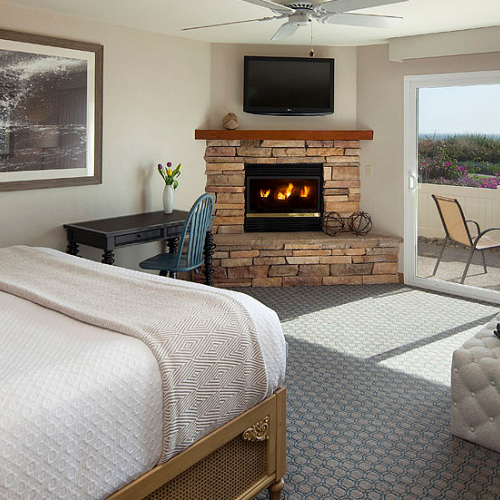A cozy hotel room with a bed, stone fireplace, wall-mounted TV, desk, and a sliding glass door leading to a balcony with outdoor seating.