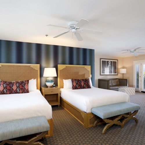 Two twin beds with wicker headboards, dark striped accent wall, nightstand with lamp, a bench at foot, and a bright, airy hotel suite.