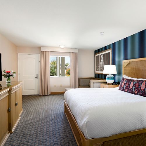 A neatly made hotel room with a large bed, a TV dresser, a blue accent wall, and a window with light curtains, ending the sentence.