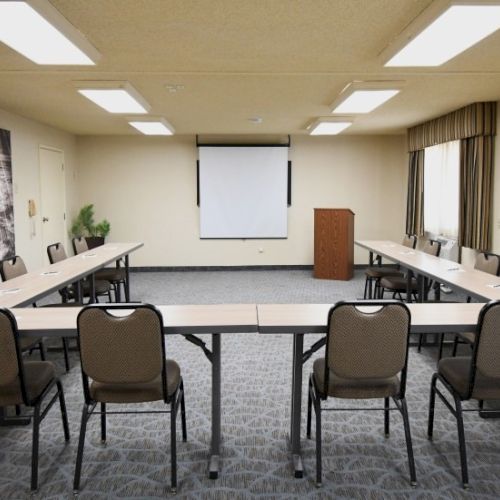 A conference room with a U-shaped table setup, beige walls, a projector screen at the front, chairs around the table, and a podium near the screen.