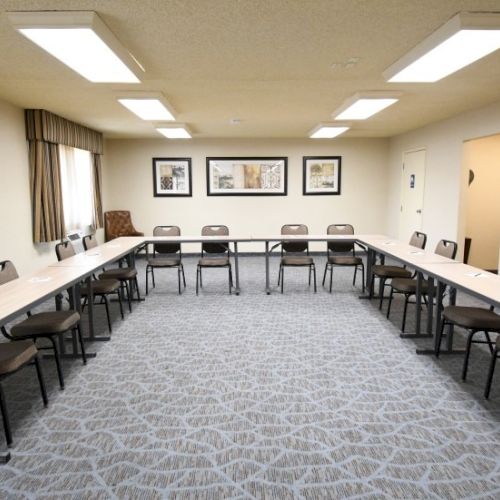 A U-shaped meeting room with long tables and chairs, cream walls, framed pictures, and overhead fluorescent lighting, carpeted floor, and a door to the right.