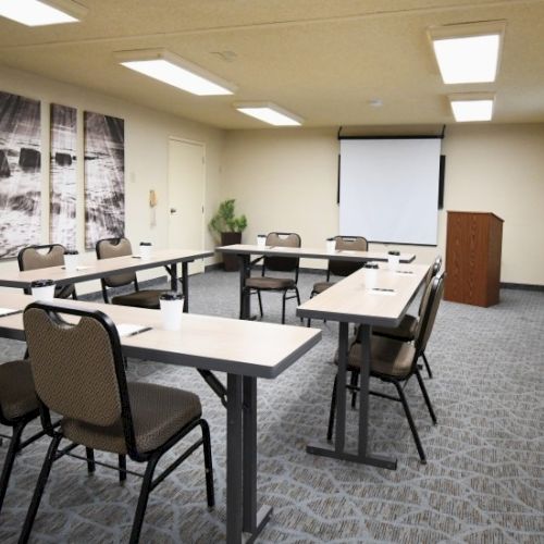 A small conference room with U-shaped desks, chairs, a projector screen, a podium, and wall art in a neutral-lit setting.