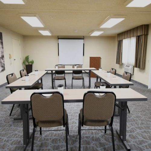 A small conference room set up for a meeting, with rectangular tables arranged in a U-shape, chairs facing a whiteboard and projector screen.