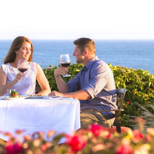 A couple sits at a seaside table for two, wine glasses raised, a bottle on the table, with the ocean and flowers in the foreground, enjoying a romantic sunset.