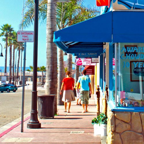 A sunny seaside street with palm trees, a blue storefront, people walking, and a sign reading &ldquo;OPEN&rdquo; in bright letters near the shop.