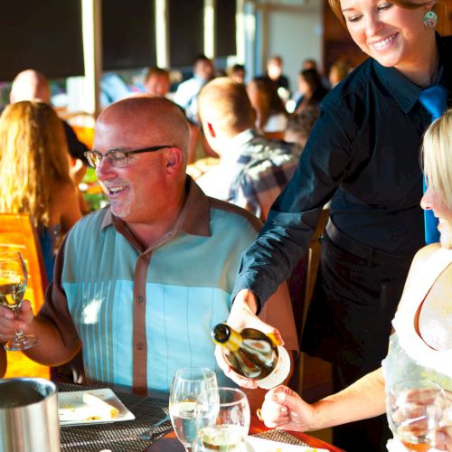 A lively restaurant scene with smiling guests, a waiter pouring wine, and people toasting at a sunlit table.