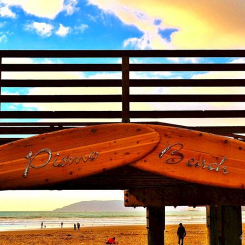 Two wooden surfboards stuck on a beach railing with a sunny sky, ocean, and people walking on the shore in the background.