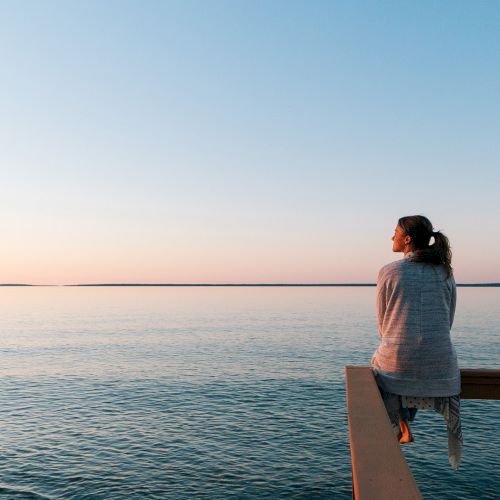 A person sits on a wooden railing by a calm body of water, watching a tranquil sunset with soft pastel skies and a distant horizon.