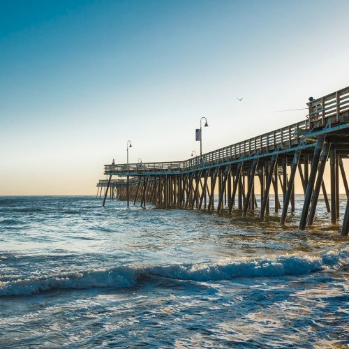 A wooden pier extends into a calm sea at sunset, with gentle waves lapping the pilings and a clear sky above, framed by warm light.