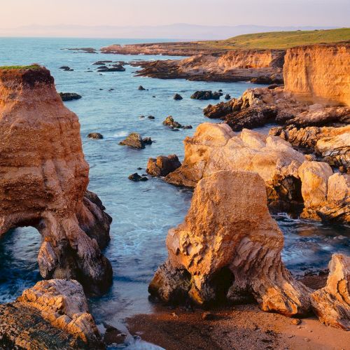 A rugged coastline with orange limestone rock formations, sea stacks, and calm blue water under a warm sunset sky.