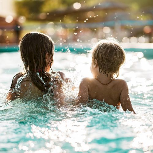 Two kids sit side by side in a sunny outdoor pool, the water sparkling as they watch the scene around them. End.