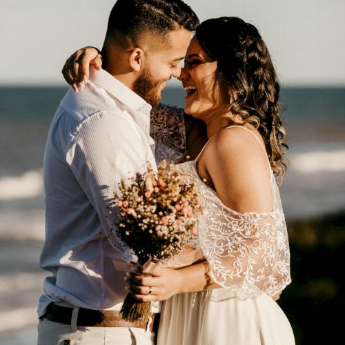 Two people embrace warmly on a beach, the man in a light shirt and the woman in a lacy dress holding a bouquet, smiling at each other.