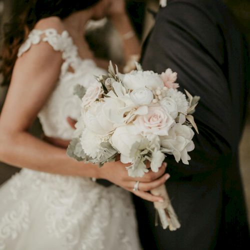 A bride in a white wedding dress holds a bouquet of pale roses, standing close to a groom in a dark suit.
