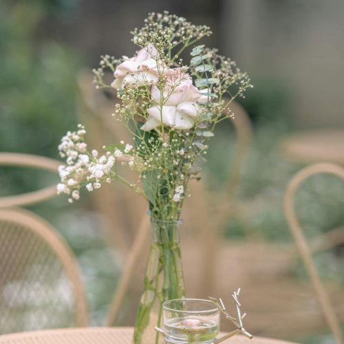 A delicate bouquet of pale pink roses with tiny white filler flowers in a slender glass vase on a wicker table, outdoors in soft daylight.