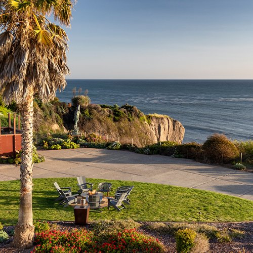 A seaside terrace with palm trees, a grassy area, outdoor seating, and a rocky cliff overlooking the ocean at sunset.