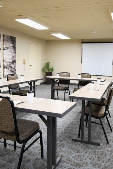 A small conference room with grey carpet, rectangular tables arranged in a U-shape, brown chairs, a projector screen, and a wooden podium.