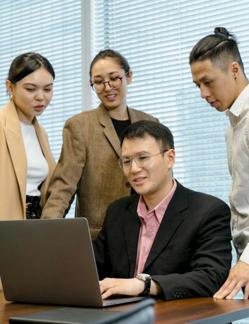 A group of four professionals gathers around a laptop in a conference room, discussing something serious and collaborating together.
