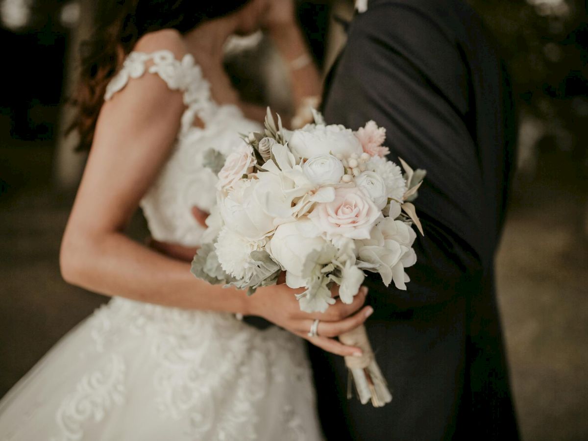 A bride in a lace wedding dress holds a pastel bouquet of roses and white flowers, standing beside a groom in a dark suit.